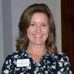 Female employee in a dark shirt with white flower wearing a name tag up against light background.