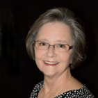 Portrait of an individual with short gray hair, glasses, and a black polka-dot top against a dark background.