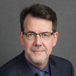 Man in dark suit wearing glasses on a dark background.