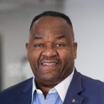 Smiling man in front of a mix of dark and light background wearing a blue jacket and a white and blue under shirt.