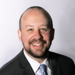 Smiling man in front of a light background wearing a dark coat, white shirt, and blue tie.