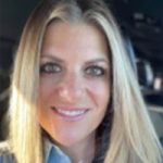 Smiling blonde woman wearing a blue collared shirt on a dark background.