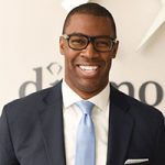 Portrait of a smiling individual in a dark suit, light blue tie, and glasses, standing in front of a wall with partial text.