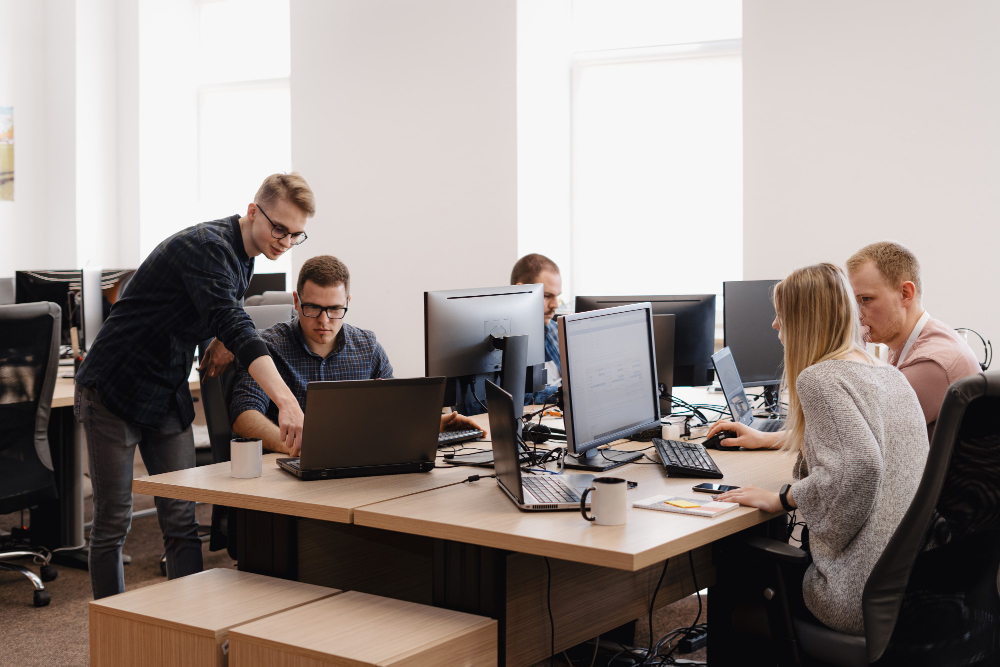 Modern office with five people working at desks equipped with computers, monitors, and coffee mugs; one person stands pointing at a laptop screen while others are seated, suggesting collaboration in a well-lit, professional workspace.