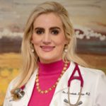Portrait of a medical professional with long blonde hair, wearing a white coat and pink top, with a stethoscope around the neck and gold-beaded necklace, standing in front of an abstract painting.